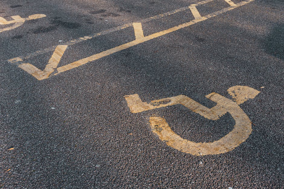 Close-up view of a black asphalt parking area with faded yellow line markings, including a symbol indicating accessible parking, along with other parking space lines. The surface shows signs of wear and weathering, with some areas of peeling paint. The image depicts a section of parking lot likely used for residential or commercial house removals, with no vehicles or moving equipment visible. It reflects the type of parking environment that may be relevant for home relocation services, such as those provided by manandvankingstonuponthames.co.uk, illustrating the importance of understanding parking and access rules during a furniture transport and loading process in Kingston upon Thames.