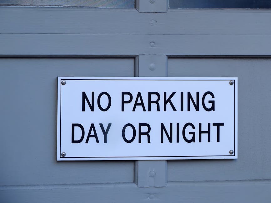 A rectangular white sign with bold black text reading 'NO PARKING DAY OR NIGHT' is mounted on a grey wooden door or wall, with four screws visible at each corner. The surface behind the sign appears smooth, and the sign is aligned centrally on the panel. The setting suggests this is an exterior location, possibly near the entrance of a property in Kingston upon Thames, which could impact parking and access for home relocation and furniture transport logistics. This image supports guidance related to parking restrictions during a house removal or moving process managed by [COMPANY_NAME], highlighting the importance of adhering to local parking rules for loading and unloading activities during the logistics of a house move, especially with considerations for access and parking rules in KT1 postcode areas.