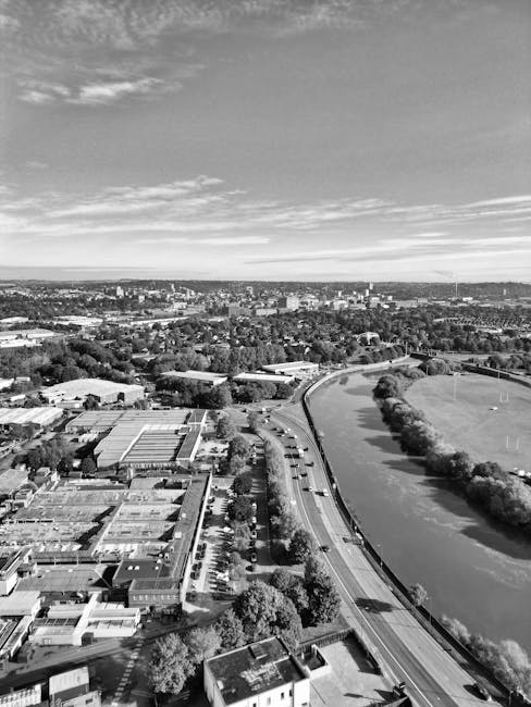 A black and white aerial photograph shows a river flowing through an urban area with trees lining its banks. To the left, there are low-rise buildings, including a large structure with a flat roof, surrounded by parking lots with several parked cars. A busy road runs parallel to the river, with vehicles visible in motion. In the background, there are more residential and commercial buildings extending towards the horizon, with a mix of green spaces and developed areas. The sky above displays scattered clouds, and the overall scene provides a view of the city's landscape from above, illustrating both natural and built environments. This image is used on the website manandvankingstonuponthames.co.uk, specifically on the page about removals in Kingston Canbury Park Road and Riverside moves, reflecting the area’s scenic views and logistical aspects of home relocation and furniture transport, captured in a clear, high-angle perspective.