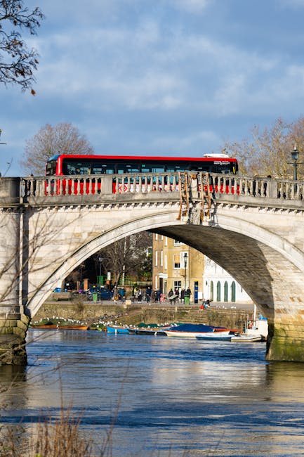 A red and white bus is crossing a stone arch bridge over a river in Kingston upon Thames, with the bridge featuring detailed balustrades and supporting stonework. Below, the river reflects the sky and surrounding trees, some of which are leafless, indicating a colder season. The background shows a row of residential buildings and trees along the riverside, with a partly cloudy sky overhead. The scene depicts an urban area with visible pedestrian pathways along the riverbank, and the bridge is positioned centrally in the image. This setting is common in house removals and relocation services in Kingston upon Thames, where careful planning involves transporting furniture, boxes, and appliances by road or across bridges during home moves. The image emphasizes the transportation logistics essential for safe and efficient moving processes, with the bus and bridge illustrating key elements of the urban environment involved in residential relocations.